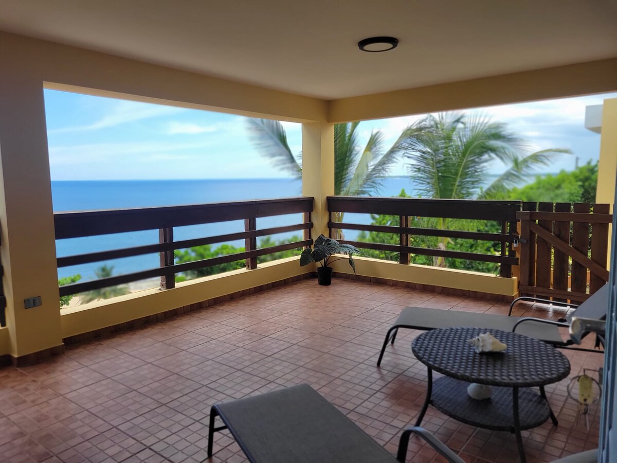 A covered terrace is presented with sweeping views of the Atlantic Ocean. Two lounge chairs and a round table are positioned on a tiled floor. Lush palm trees frame the scene, adding to the relaxing ambiance of the space.