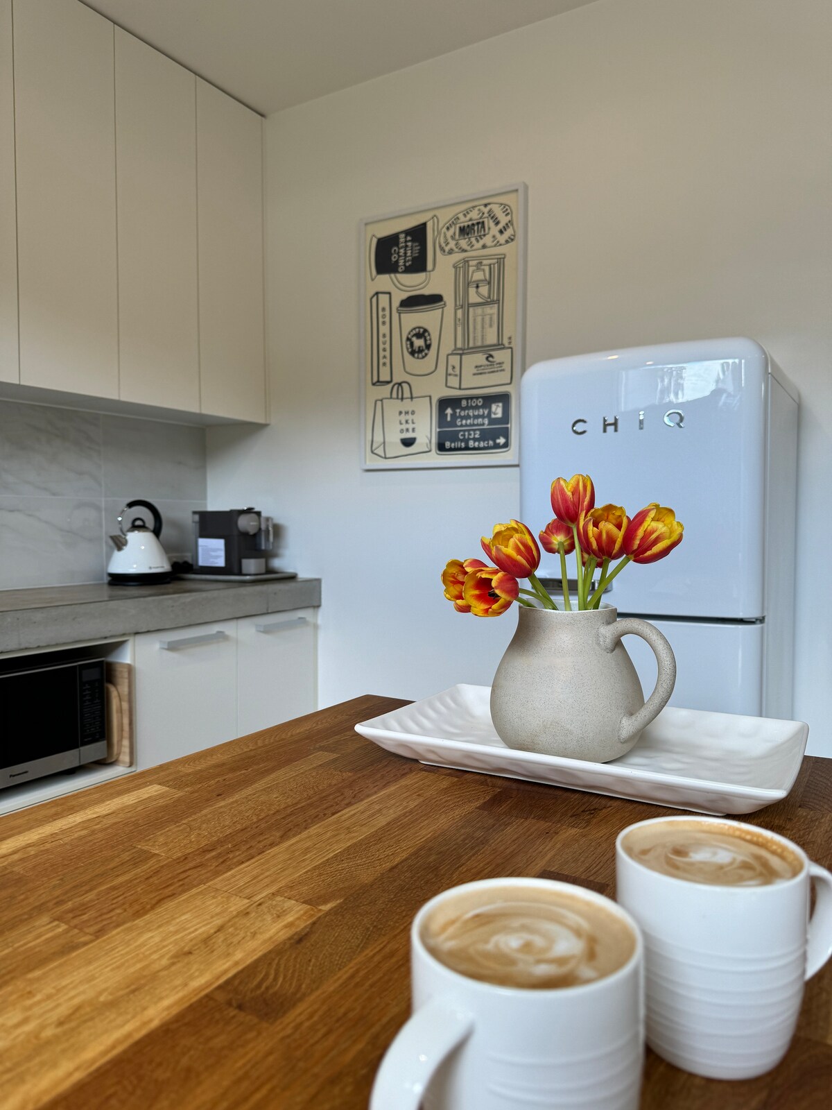 A bright kitchenette features a white refrigerator, wooden countertop, and stylish cabinetry. A neutral vase filled with vibrant tulips adds a pop of color. Two cups of coffee are placed on a simple tray, creating a welcoming and functional space for enjoying warm drinks.