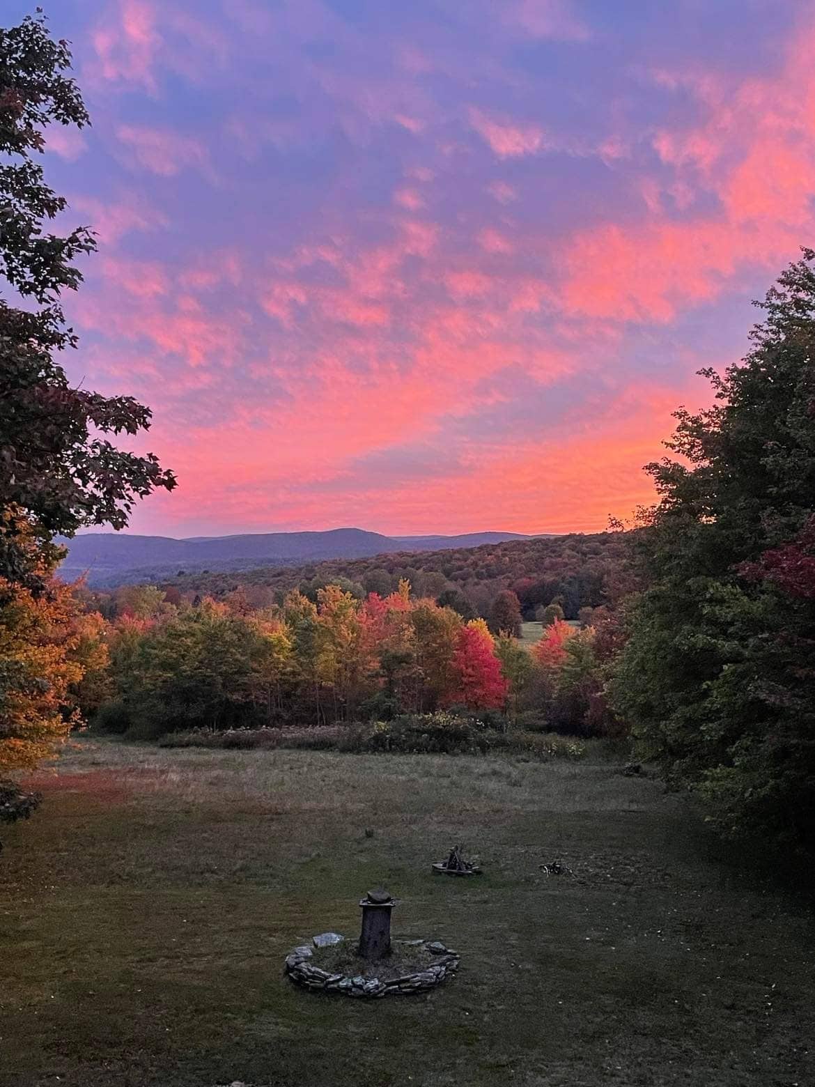 A colorful sunset paints the sky in shades of pink and purple, casting a warm glow over the landscape. The foreground features a simple stone fire pit surrounded by grassy areas, framed by trees with autumn foliage in various hues.