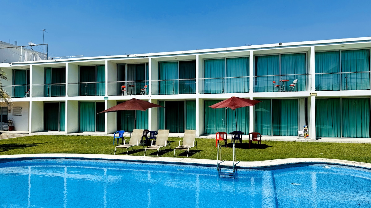 A serene view of a blue swimming pool in the foreground, bordered by green grass. Behind the pool, a modern two-story building with multiple balconies is visible, featuring large glass doors and flowing turquoise curtains. Lounge chairs and umbrellas are arranged in a relaxed outdoor setting.