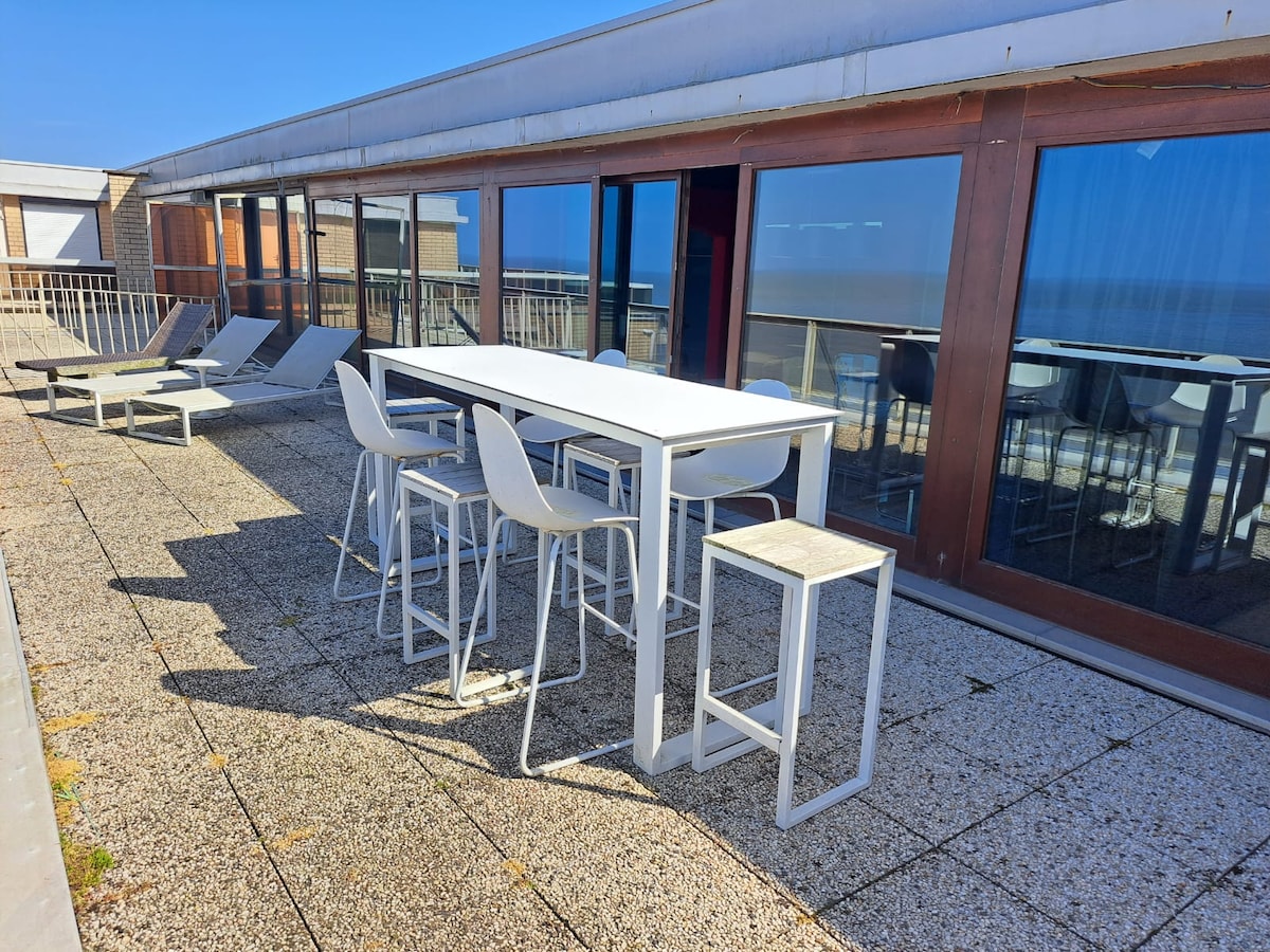 A spacious terrace is presented, featuring a white outdoor dining table surrounded by white chairs. Multiple lounge chairs are positioned nearby, all overlooking the sea. The surface is finished with stone tiles, and large windows from the penthouse reflect the clear blue sky.