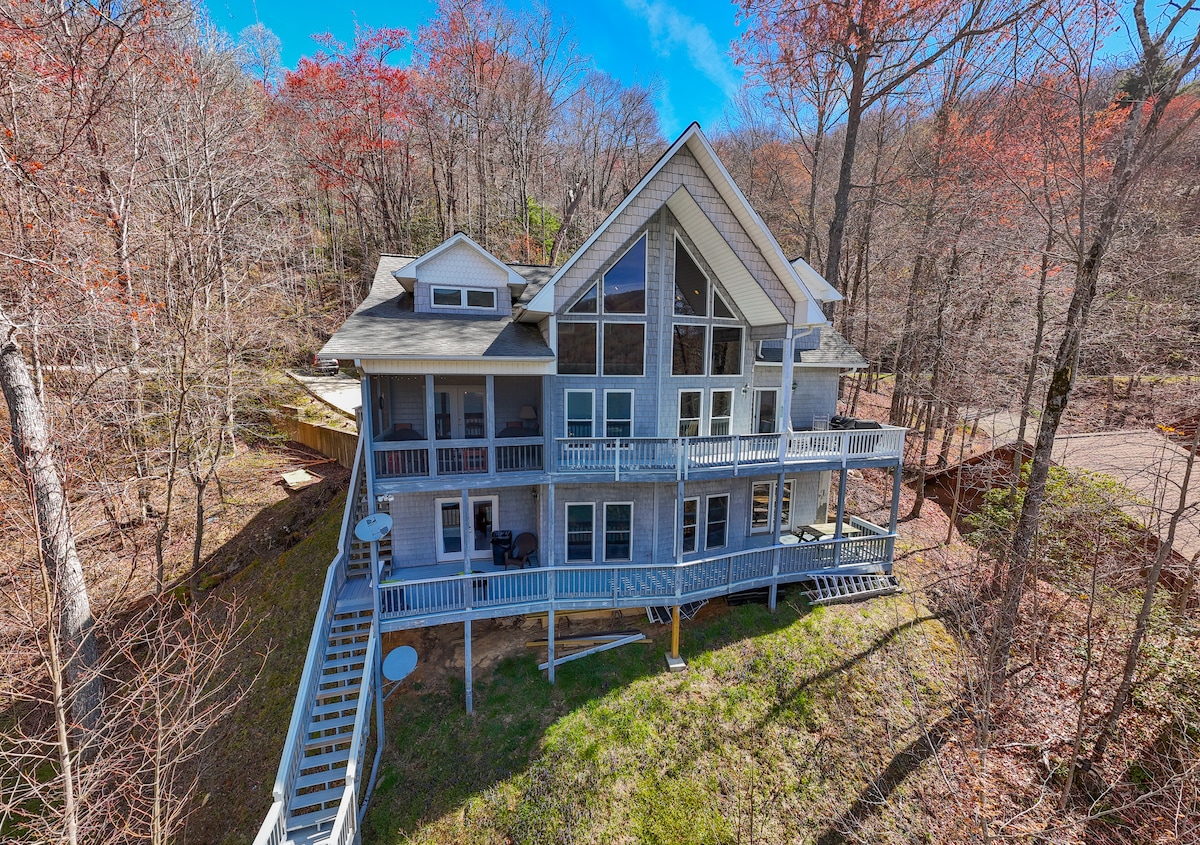 The lakefront home features multiple levels with large windows, showcasing the natural surroundings. A spacious deck wraps around the house, providing outdoor seating areas. The structure is set against a backdrop of trees, highlighting the serene and secluded location near Lake Nantahala.
