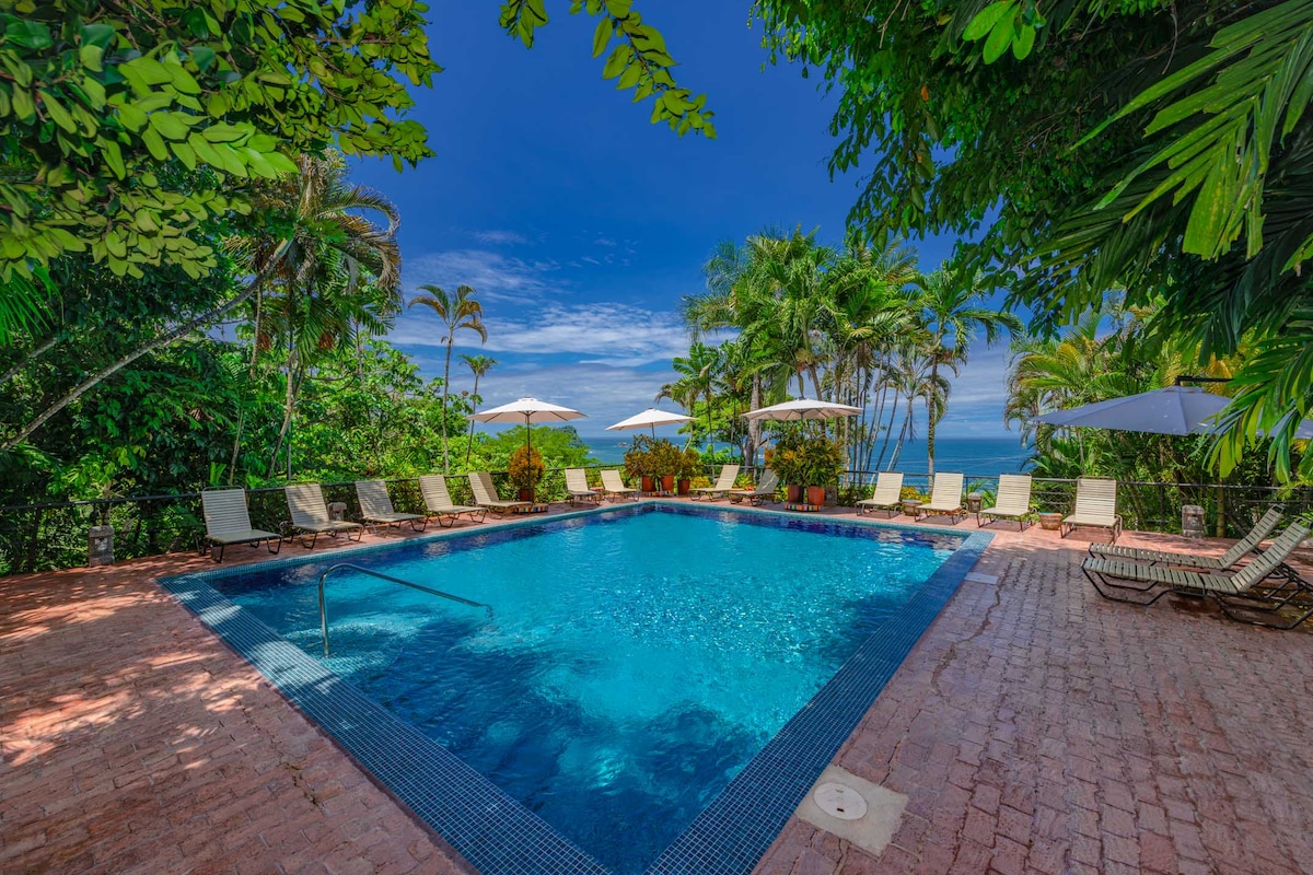 A large swimming pool is surrounded by a brick patio and lush greenery. Sun loungers with umbrellas are positioned around the pool, offering shaded relaxation. The vibrant blue water of the pool contrasts with the serene sky and ocean visible in the background.