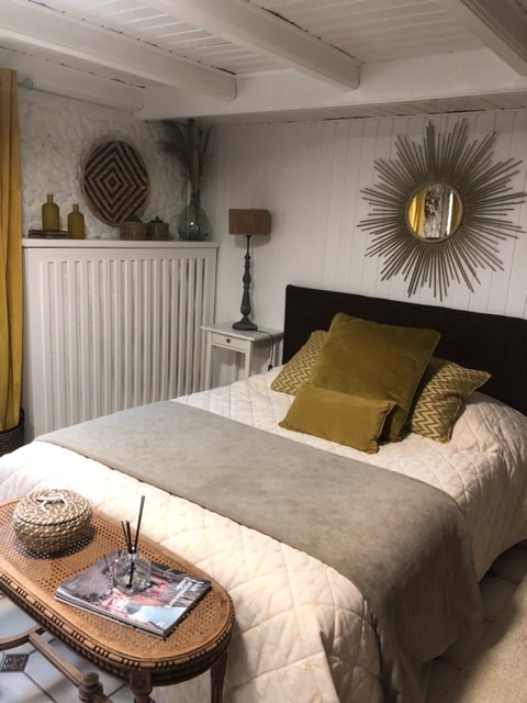 A cozy sleeping area is presented with a spacious bed, adorned with soft, neutral linens and a mix of decorative pillows. A low coffee table sits in front of the bed, featuring a magazine and a decorative piece. Natural light enters through a small window.