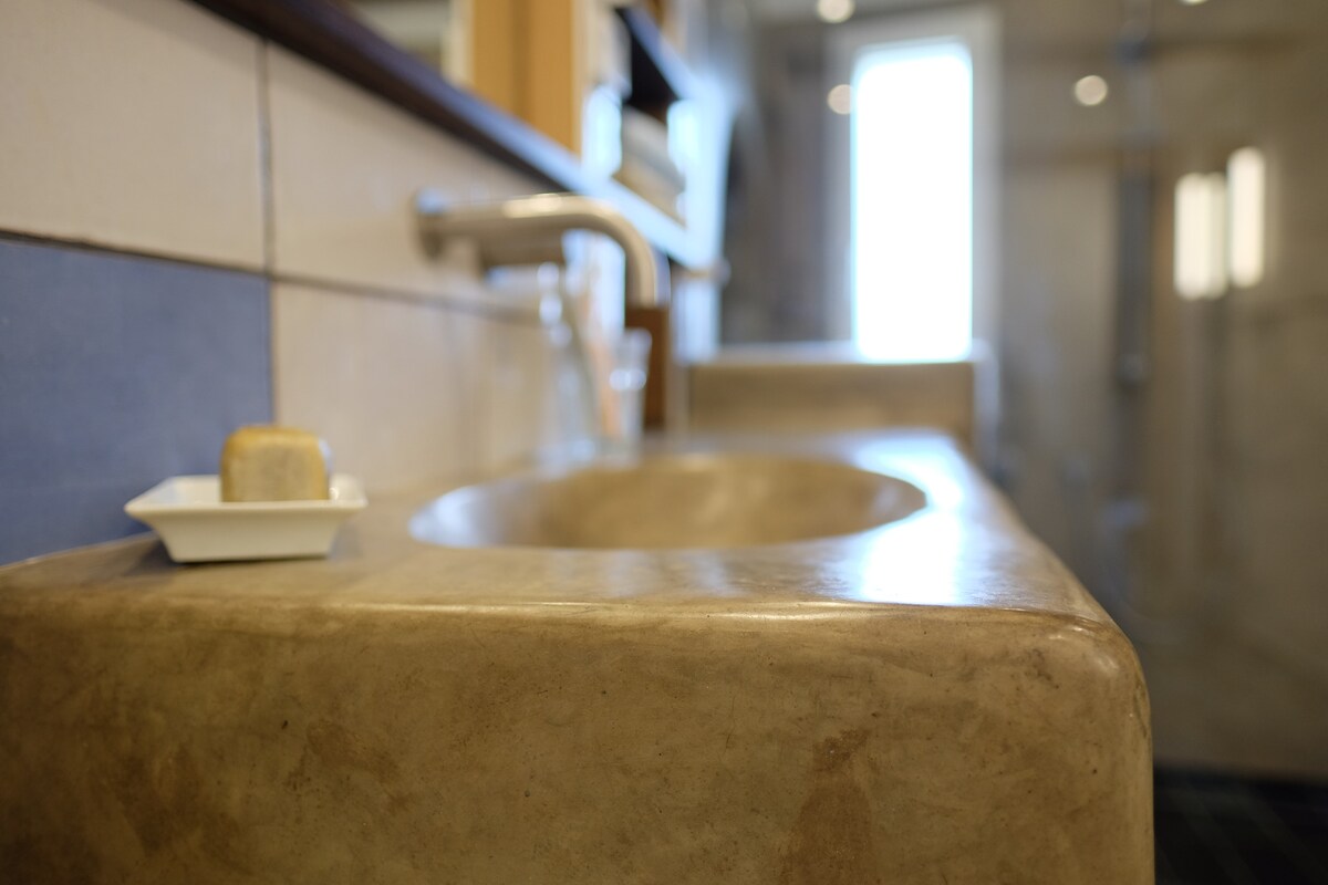 The bathroom features a unique vessel sink crafted from tadelakt finish, complemented by a wall-mounted faucet. Natural light enters through a nearby window, illuminating the space. A small dish with a soap bar rests beside the sink, highlighting the thoughtful design.