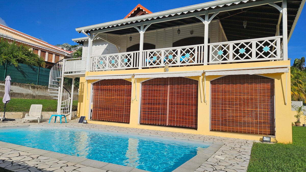 A spacious villa is seen from the poolside, featuring a charming balcony with wooden railings and a vibrant yellow exterior. The inviting pool reflects clear blue skies, and decorative bamboo shades cover large windows. Lush greenery frames the property, creating a tranquil outdoor setting.