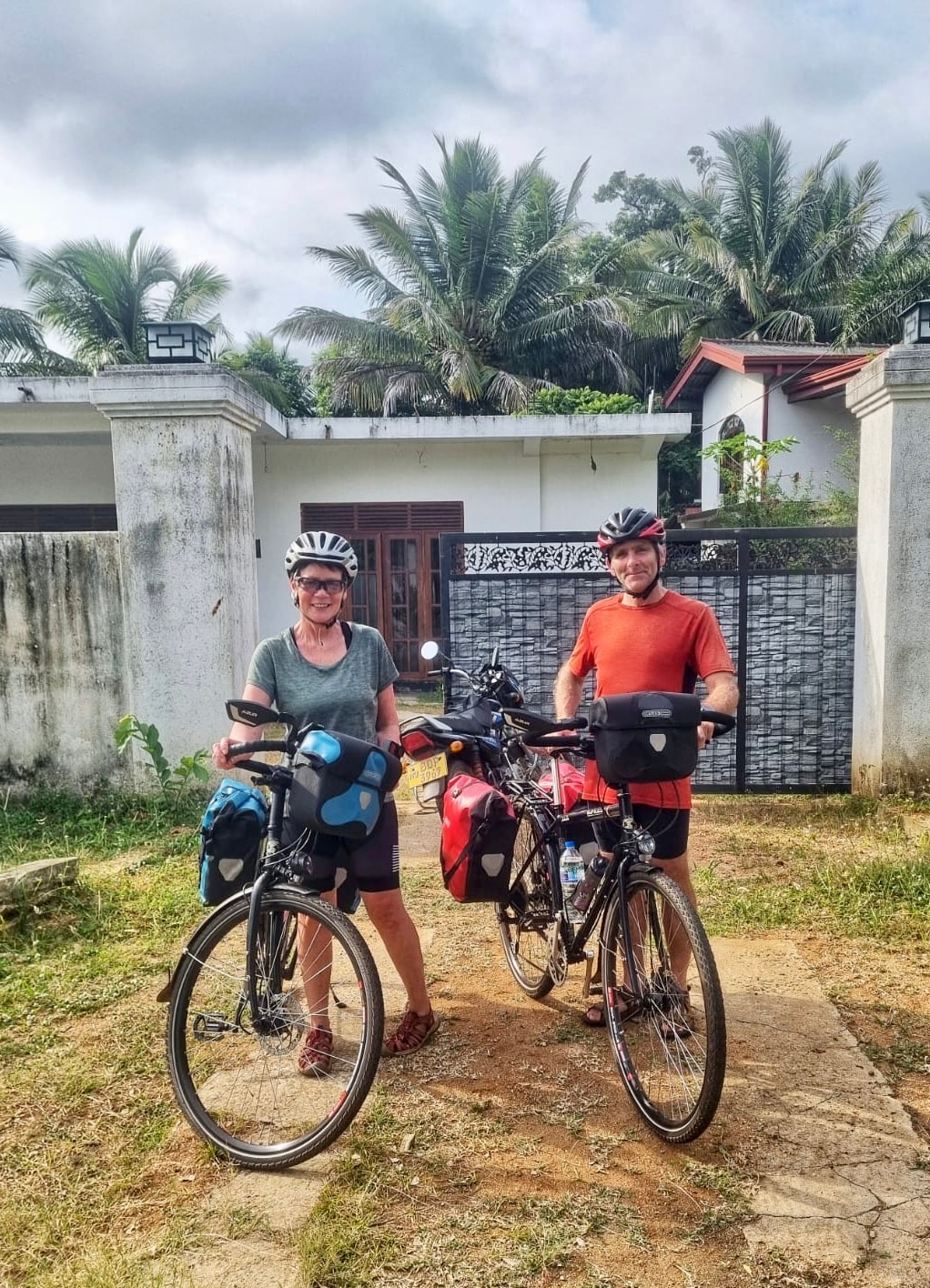 Two cyclists are positioned in front of a house surrounded by a lush garden. Each bicycle is equipped with panniers, indicating preparation for travel. The scene is framed by palm trees and a partial view of the house's entrance, highlighting the outdoor setting.