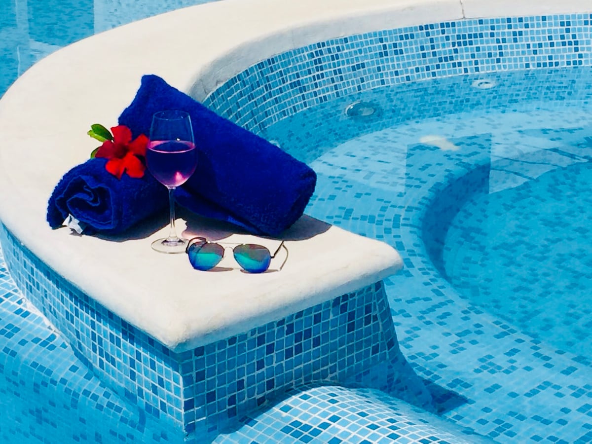 A shaded ledge beside a sparkling jacuzzi is adorned with a blue towel, a glass of pink beverage, and a pair of sunglasses. The vibrant tile pattern and clear blue water create a refreshing atmosphere.
