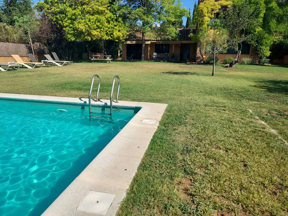 A private pool is framed by a lush green garden, with steps leading down to the water's edge. Sun loungers are positioned on the grass, and a spacious patio area can be seen in the background, surrounded by trees and natural foliage.