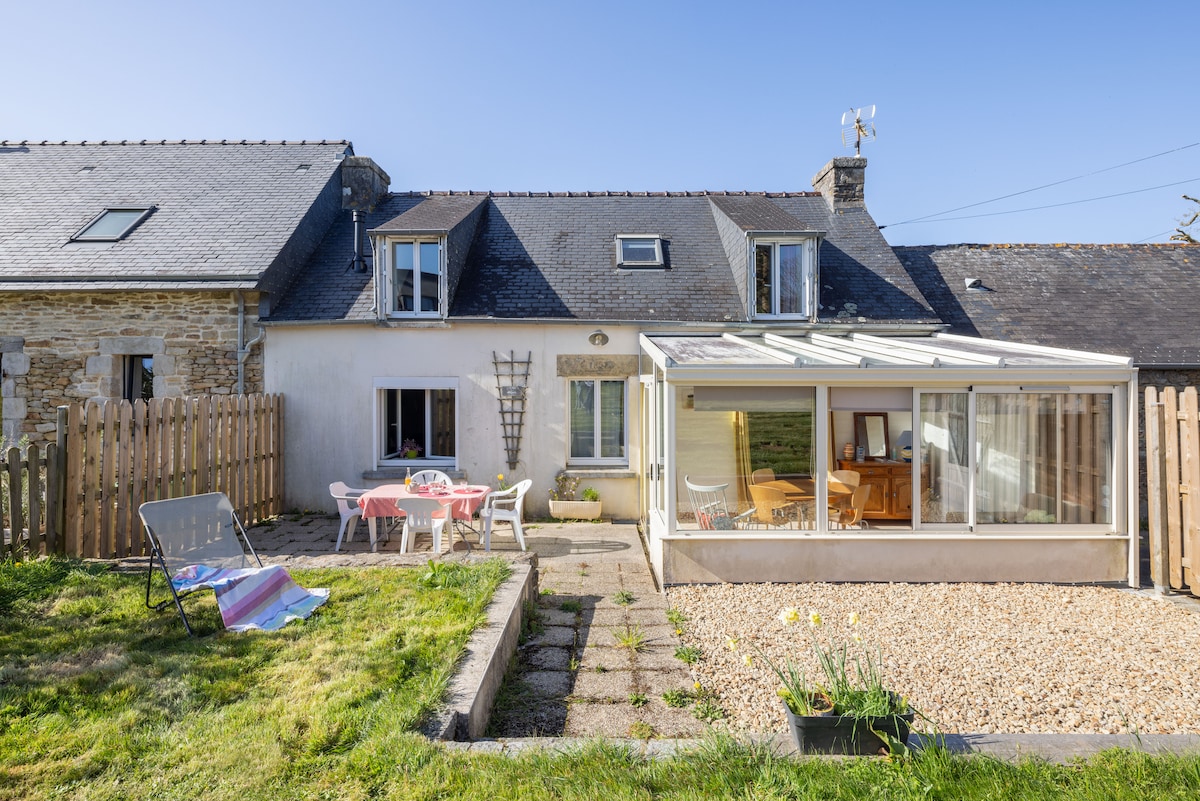 A spacious outdoor area is displayed, featuring a sunlit terrace with a glass conservatory connected to the house. A dining table is set for gatherings, surrounded by light-colored chairs. A lawn with a striped blanket and a lounge chair invites relaxation amidst a peaceful setting.