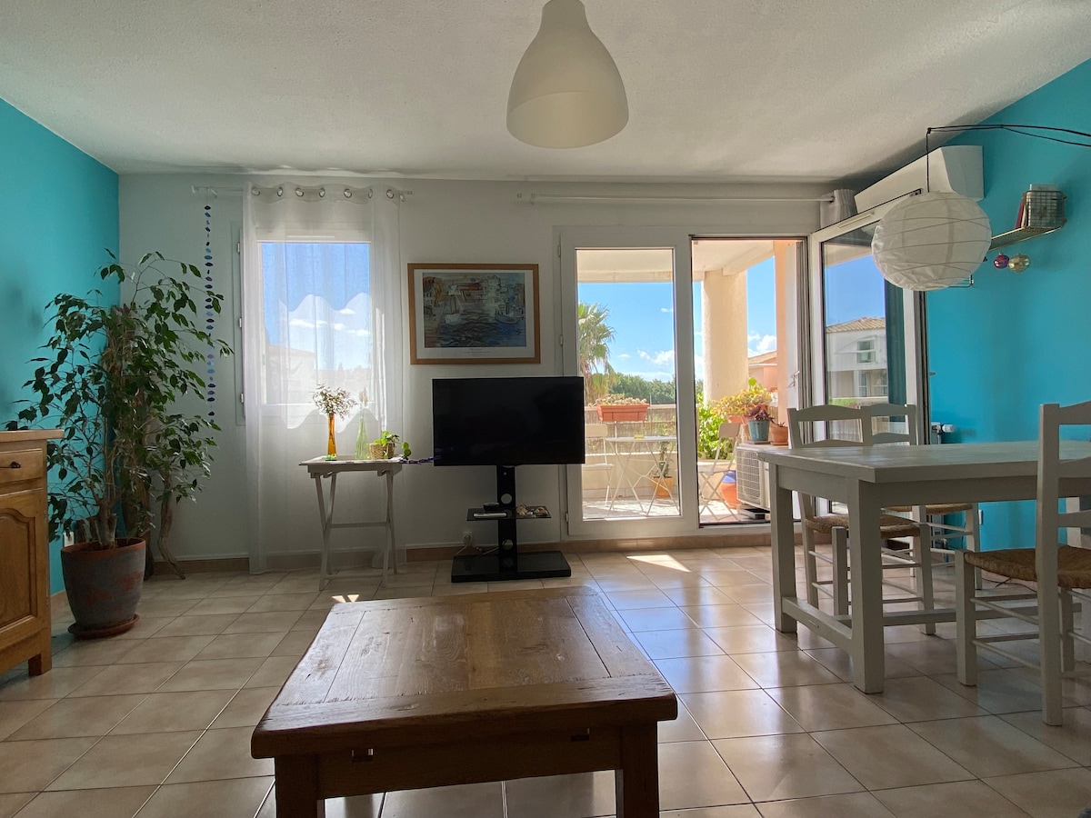 A bright living area features a turquoise accent wall and large sliding glass doors, allowing natural light to fill the space. A wooden dining table and white chairs are positioned nearby. Indoor plants add greenery, with a television mounted on a stand in the corner.