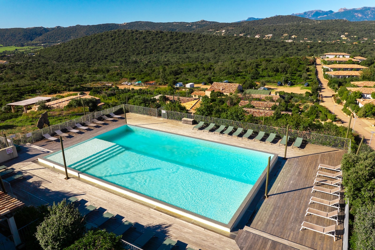 An expansive outdoor mirror pool is set against a backdrop of rolling green hills. Sun loungers line the wooden deck, providing a spacious area for relaxation. The clear water reflects the surrounding landscape, creating a serene and inviting space.