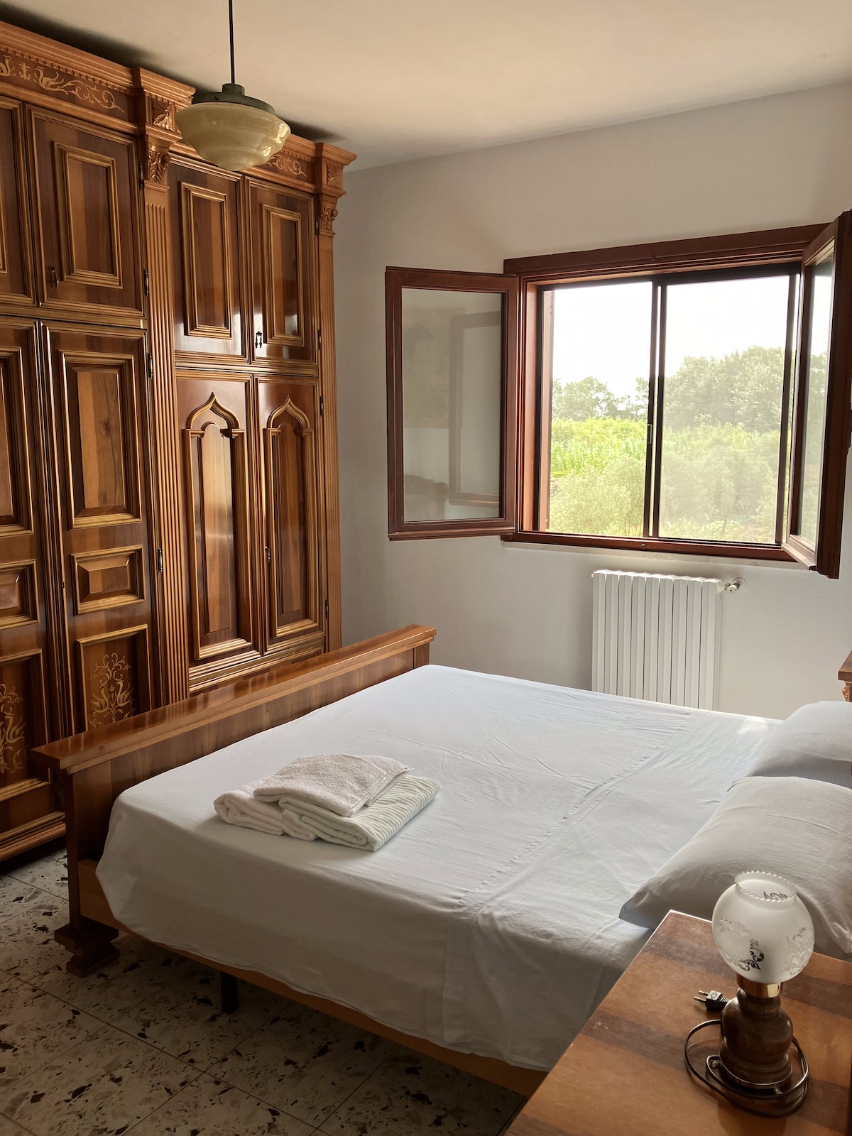 A serene bedroom features a spacious bed dressed in crisp white linens, accompanied by neatly folded towels. A wooden wardrobe with intricate designs adds warmth, while the large window allows natural light to fill the room. Views of greenery are visible outside.