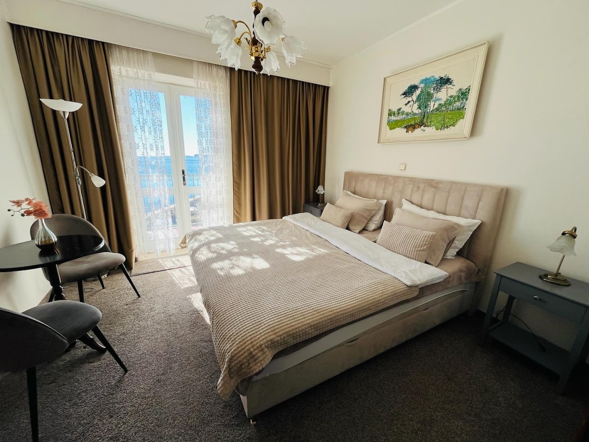 A serene bedroom features a spacious bed dressed in light textiles, complemented by soft pillows. Tall windows with sheer curtains allow natural light to fill the room, while a small table and chairs provide a cozy seating area. An artwork depicting a landscape is mounted above the bed.