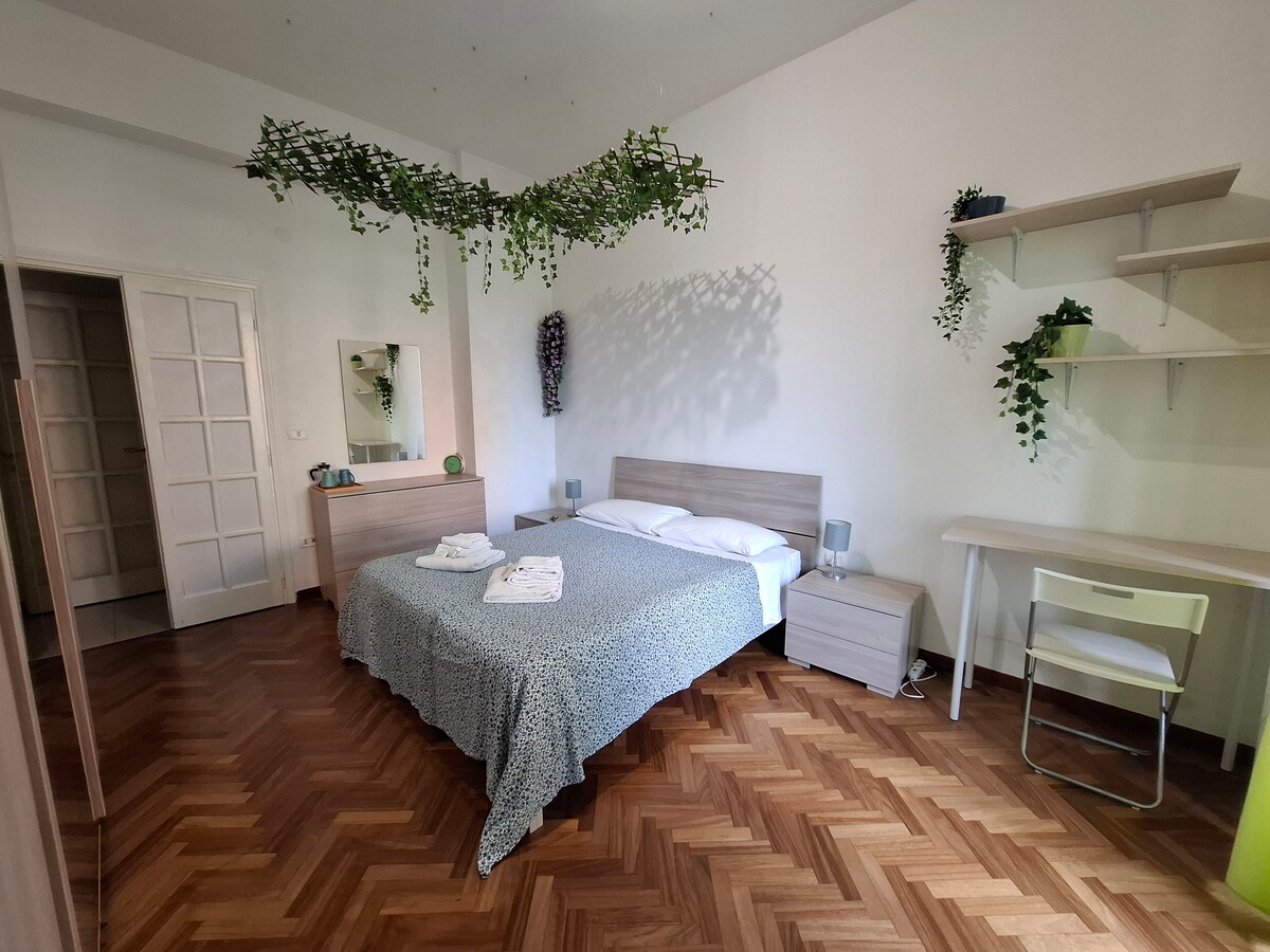 A spacious bedroom features a double bed with a patterned blanket and decorative bed linens. Two bedside tables with lamps flank the bed. A work desk and chair are positioned against the wall. Plants are displayed on shelves and hang from the ceiling, enhancing the room's character.