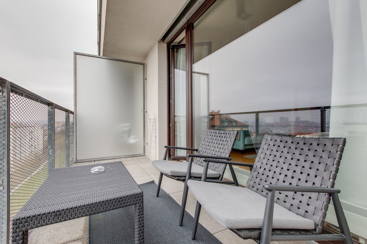 An outdoor terrace is equipped with two grey chairs and a small, matching table. Glass doors provide access to the interior, while a view of the surrounding area is visible through the protective railing. A neutral rug enhances the outdoor seating area.