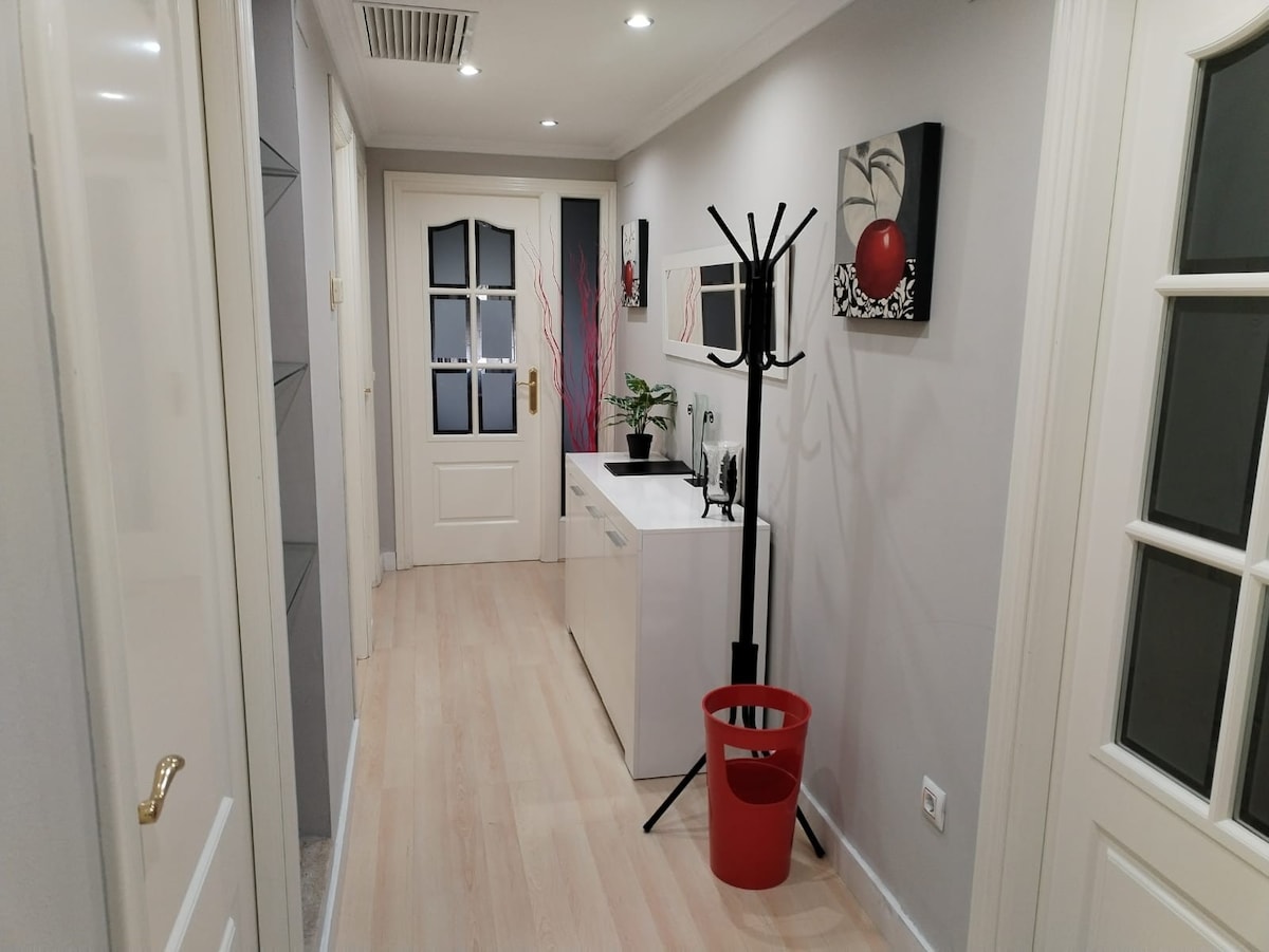 A well-lit hallway features light-colored wooden flooring and pale walls adorned with modern art. A coat rack stands beside a white cabinet, which is complemented by a small plant. A red bin adds a pop of color to the space, creating a neat and organized appearance.