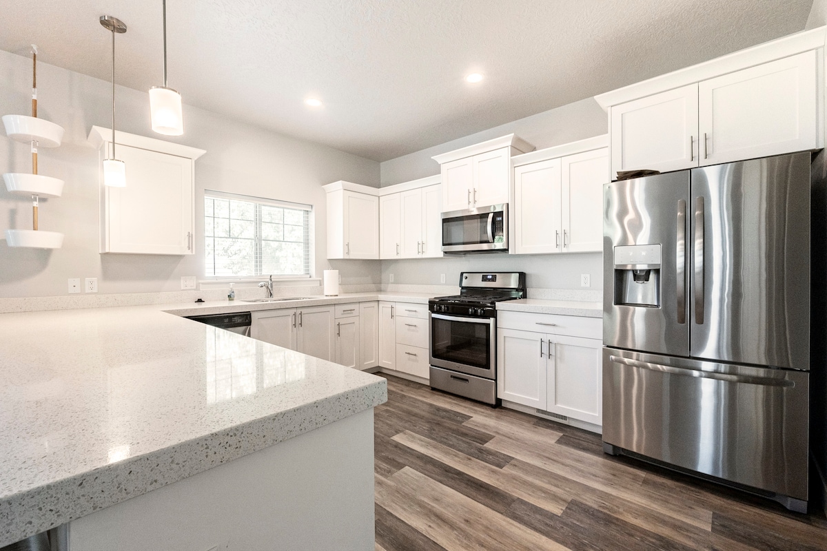 A modern kitchen features sleek white cabinetry and stainless steel appliances. Light countertops provide a spacious preparation area. A window allows natural light to fill the room, with warm wooden flooring contrasting the minimalist design.