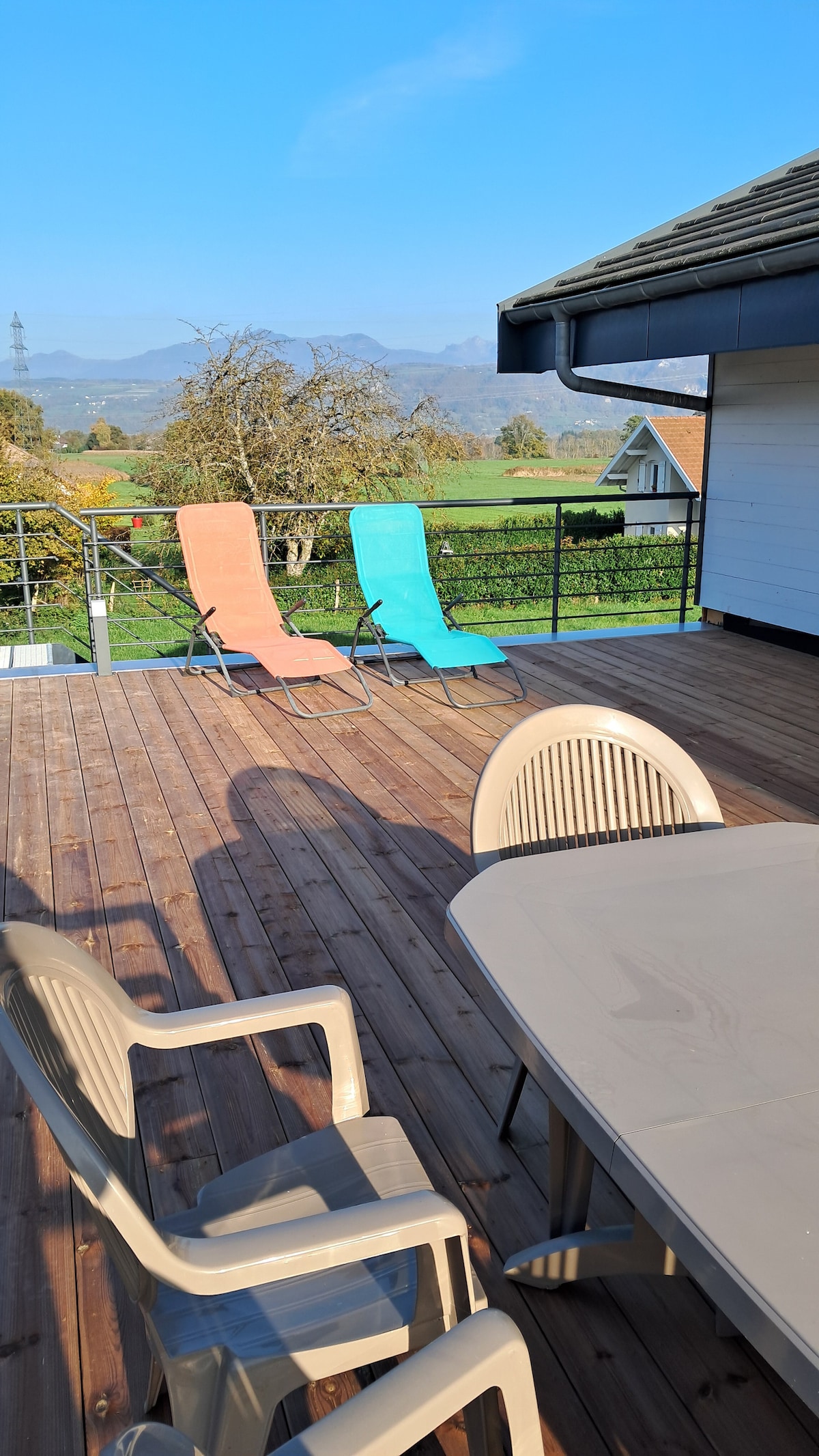 A spacious terrace features a wooden floor and furniture, including a round table with multiple chairs. Two brightly colored lounge chairs are positioned under clear blue skies, offering views of the surrounding green landscape and distant mountains.