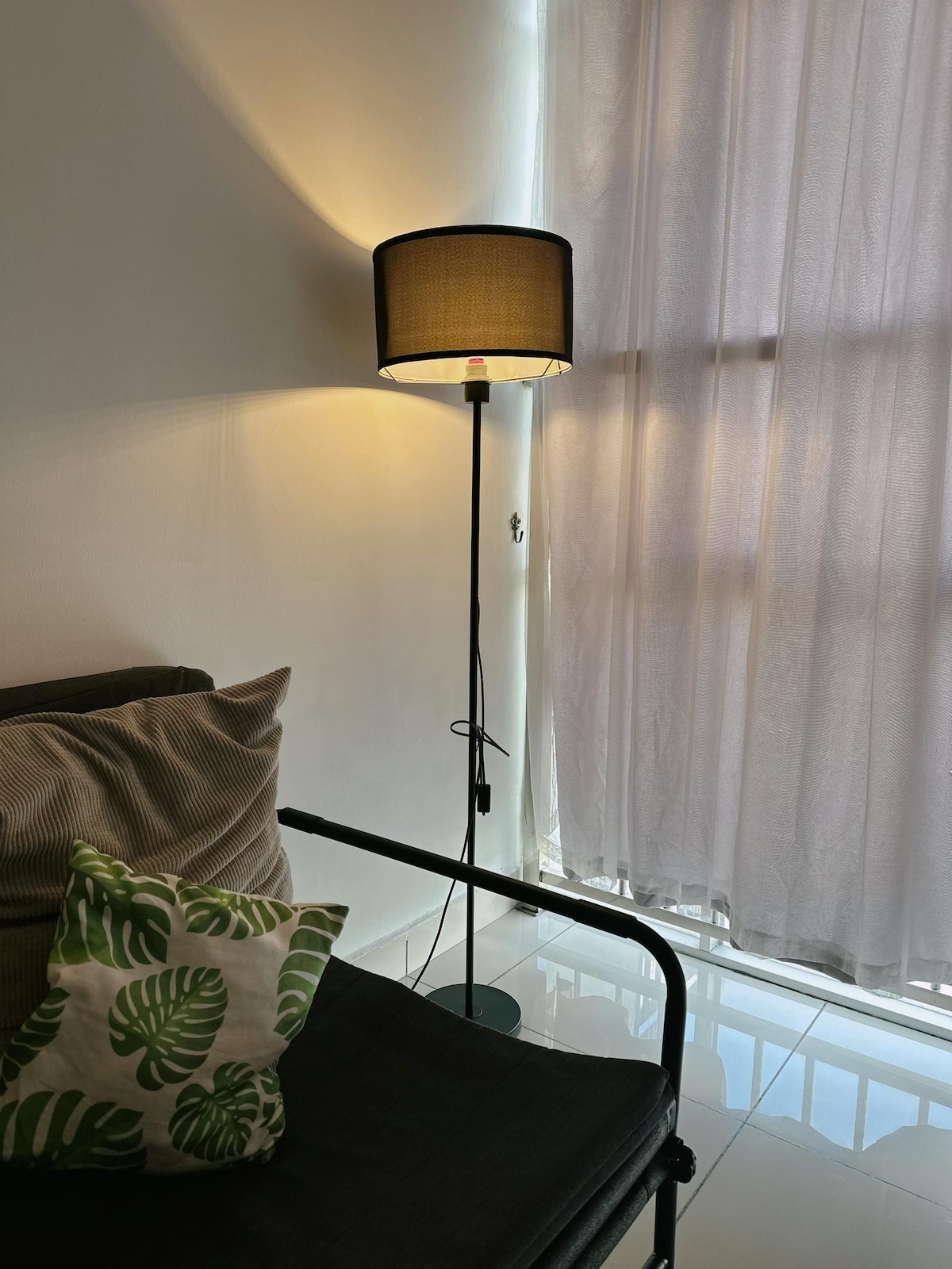 A floor lamp with a black shade is positioned next to a black metal chair. The light emanates a soft glow, highlighting the nearby window covered with sheer curtains. A decorative pillow with a leafy pattern adds a touch of color to the seating area.