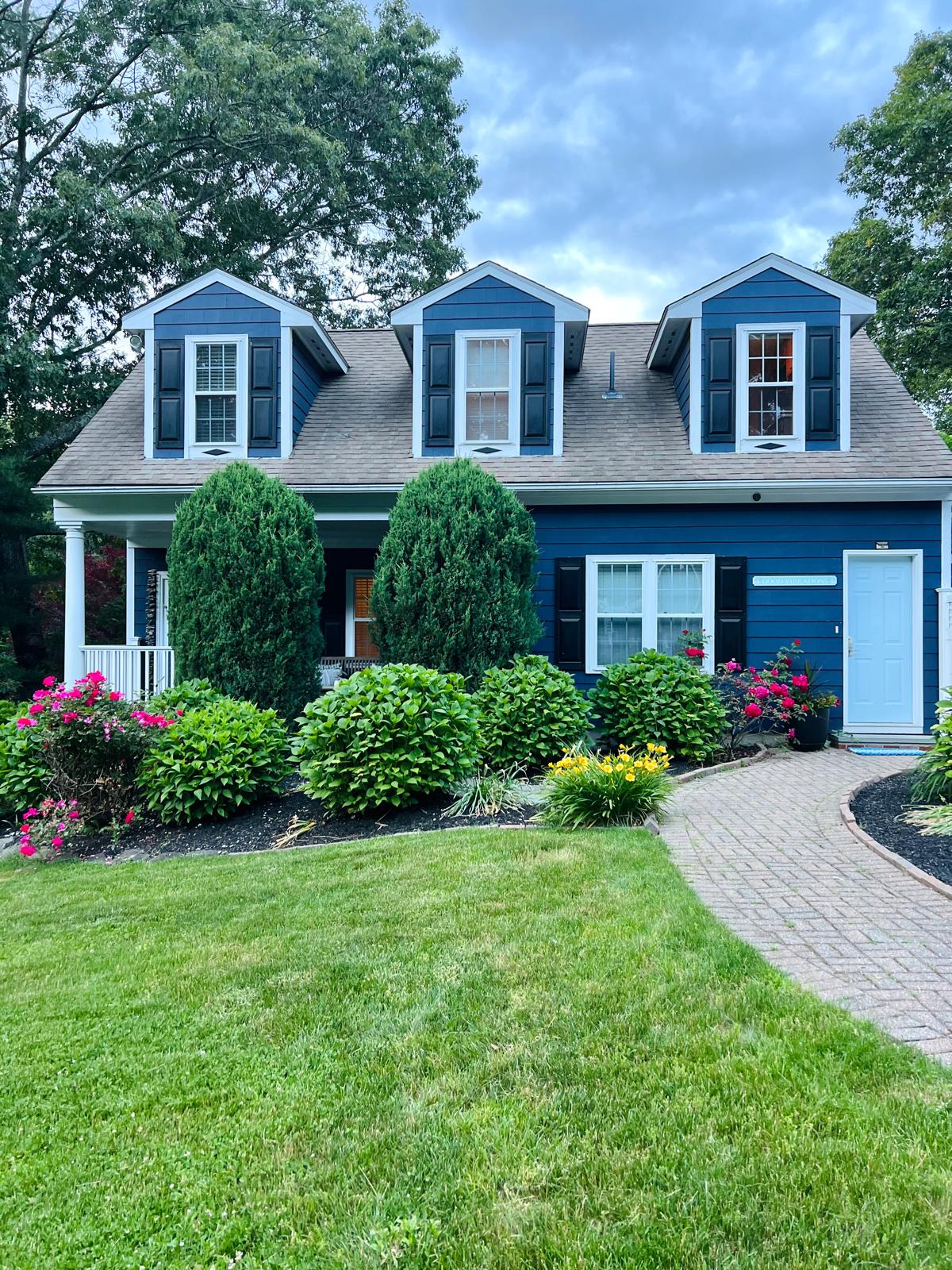 A charming blue guest house features a welcoming front entrance with a light blue door. Lush green shrubs and colorful flower beds surround the pathway, leading to a covered porch. Three dormer windows provide additional character to the inviting exterior.
