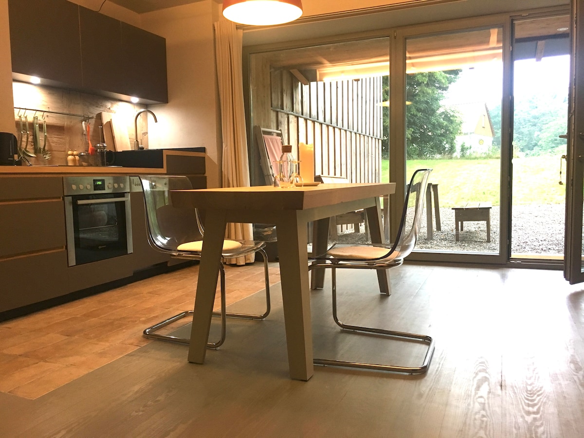A modern kitchen area is visible, featuring a sleek countertop and a dining table with clear chairs. Large sliding glass doors open to the outdoors, allowing natural light and views of the surrounding greenery. The space is designed with neutral tones and minimalist decor.