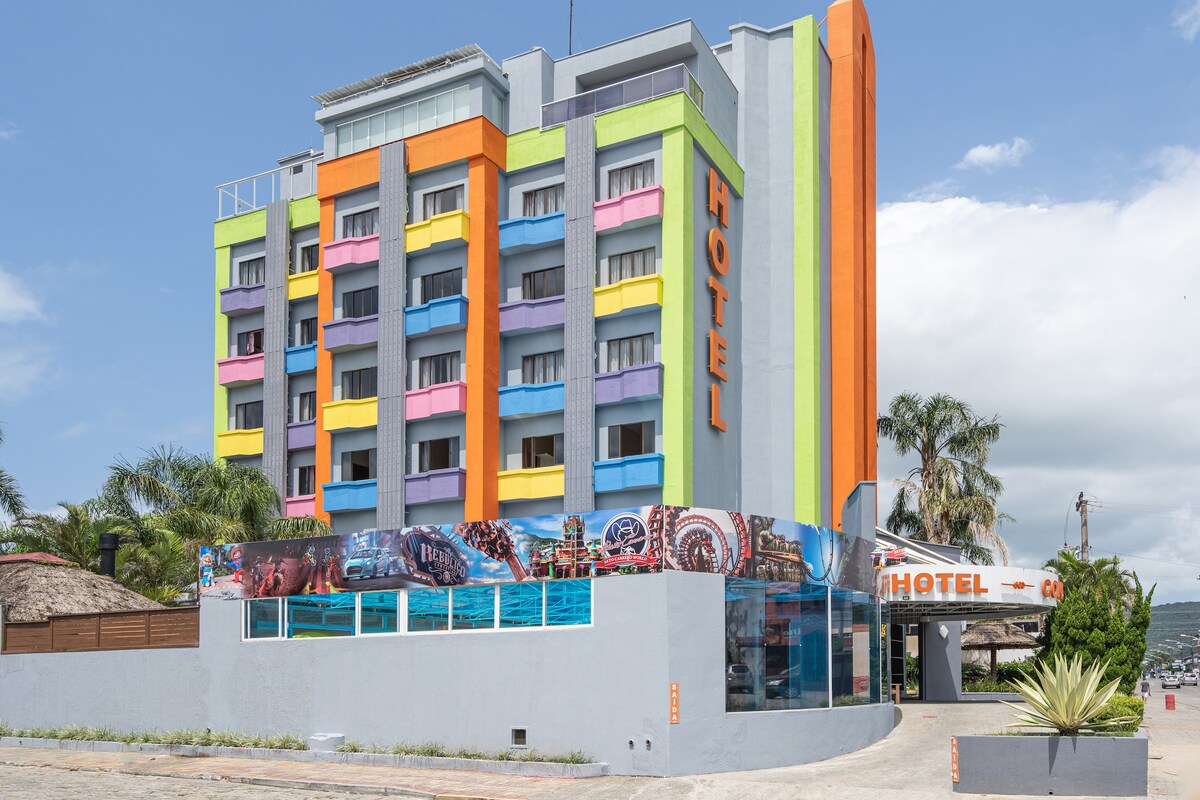 The hotel exterior presents a modern multi-story building featuring a colorful facade with vertical stripes in bright hues. Large windows allow natural light to enter, while a driveway leads to the entrance, surrounded by tropical landscaping.