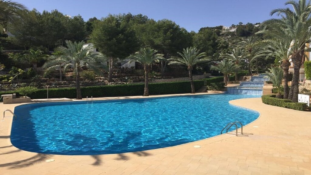 A large swimming pool is surrounded by palm trees, providing a tranquil setting. The clear blue water glistens in the sunlight, and a gently sloping pathway leads to a smaller pool. Lush greenery and terraced landscaping create a peaceful environment.