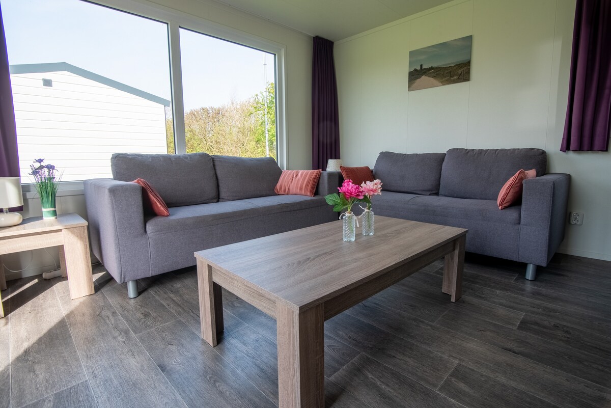 The living area features two comfortable gray sofas adorned with colorful cushions. A wooden coffee table sits centrally, complemented by vases containing fresh flowers. Large windows allow natural light to fill the space, framed by dark curtains.