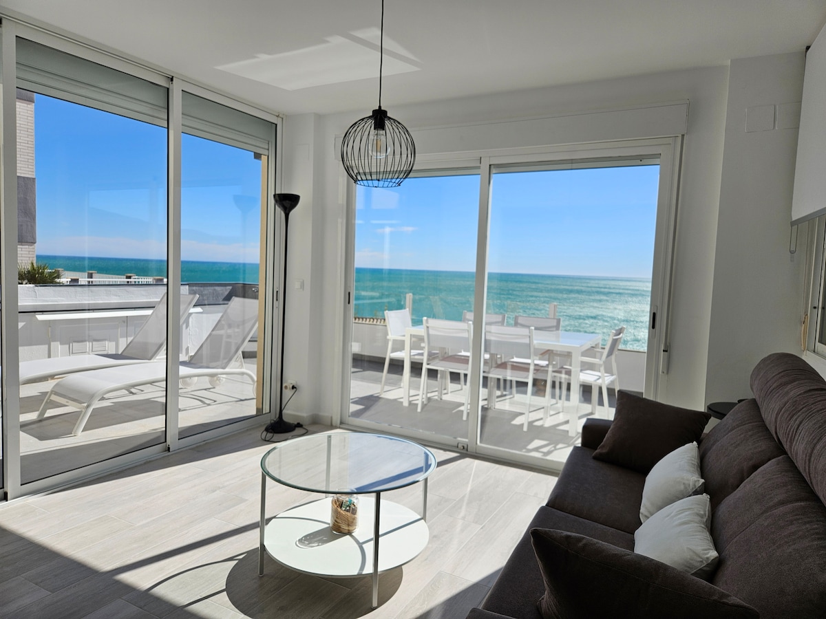 A bright living area features expansive glass doors that open to a terrace overlooking the sea. Natural light fills the space, highlighting a comfortable dark sofa and a round glass coffee table. Outdoor furniture can be seen on the terrace, perfect for enjoying views.