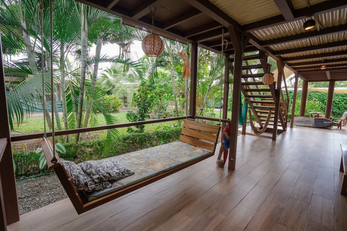 A welcoming porch area features a large wooden swing bed adorned with a patterned cushion. Lush greenery surrounds the space, with tropical plants visible through expansive windows. A spiral staircase leads to an upper level, and a cozy seating area is positioned nearby.