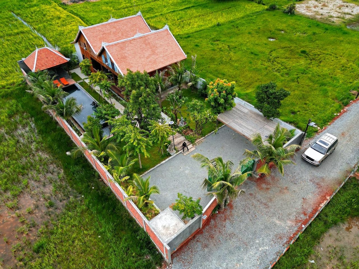 An aerial view of a private villa surrounded by lush greenery features a red-tiled roof and landscaped gardens. A private swimming pool is visible beside the villa, with palm trees adding to the serene setting. A vehicle is parked on a gravel path leading to the entrance.