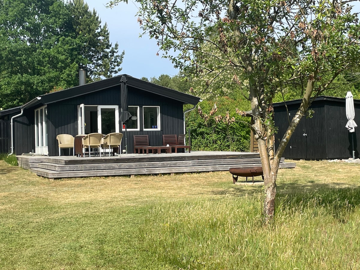 A black wooden summer house sits on a spacious lawn with a wooden deck extending from it. Outdoor seating arrangements, including chairs and a table, are visible. A tree and a parasol contribute to the outdoor setting, framed by lush greenery.