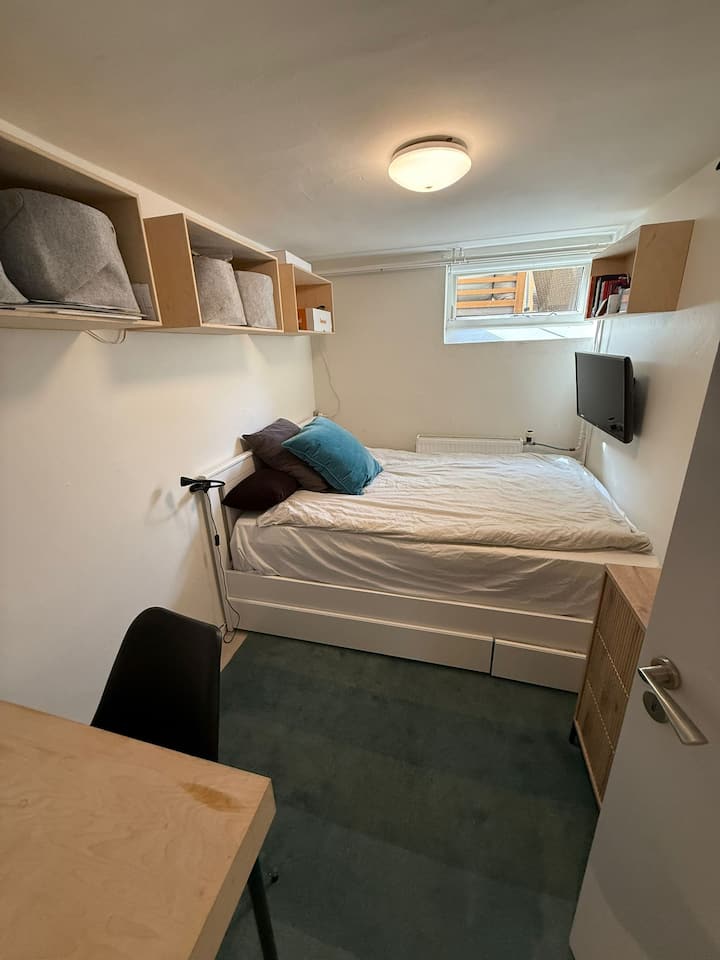 Útulná spálňa v suteréne s manželskou posteľou, televízorom, stolom a zásuvkami na oblečenie.