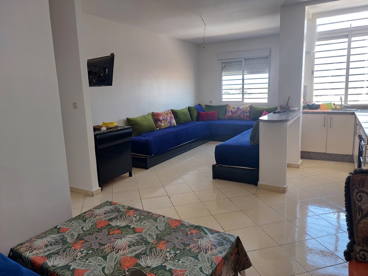 A spacious living area is presented with a large blue sectional sofa adorned with colorful cushions. A small dining table with a patterned tablecloth sits in the foreground, while natural light floods the room through large windows, enhancing the airy atmosphere.