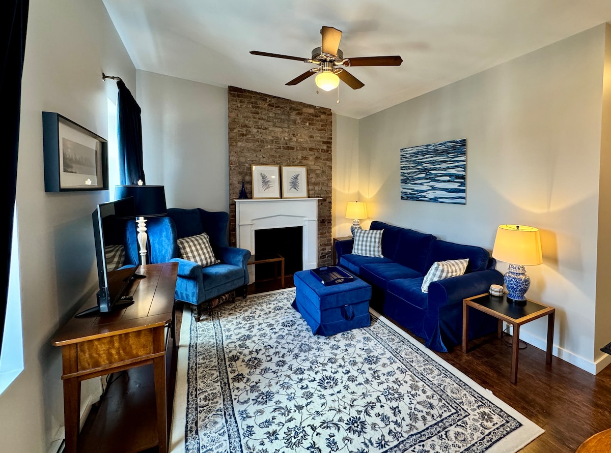 The living room features a comfortable blue sofa and matching chairs, arranged around a central ottoman. A patterned area rug covers the floor, while a ceiling fan provides overhead lighting. Warm lamps flank the seating area, and a decorative fireplace offers a charming focal point.