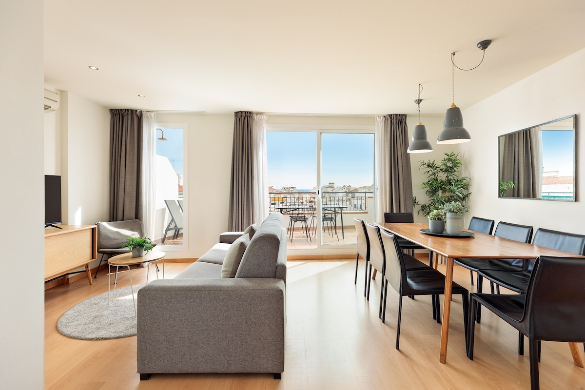 A spacious living area features a comfortable sectional sofa and a dining table with dark chairs. Large windows allow natural light to illuminate the room, while a balcony provides views of the surrounding area. Decorative elements include a potted plant and wall-mounted mirrors.