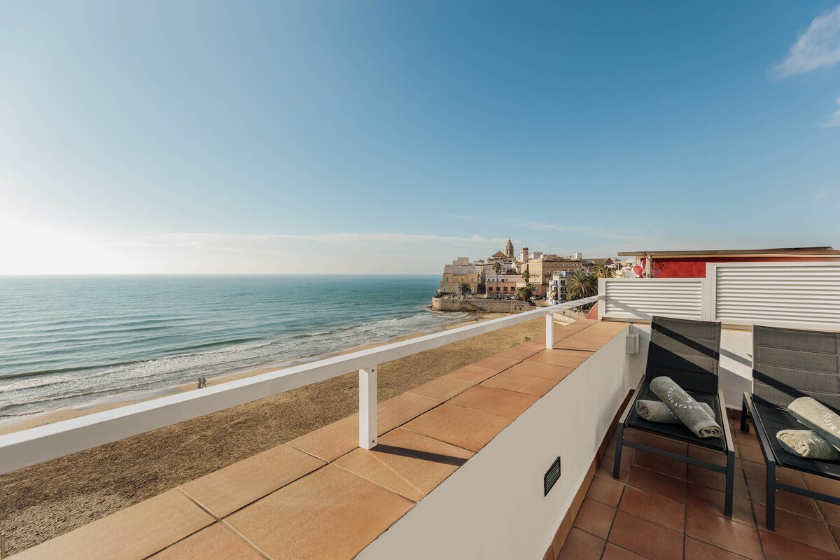 A spacious terrace overlooks the beach, featuring two lounge chairs and a beautiful view of the ocean. The coastline and distant buildings are visible under a clear blue sky, creating a serene setting for relaxation.