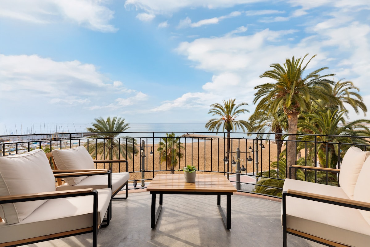 A spacious terrace features two armchairs and a coffee table overlooking the Mediterranean Sea and white sand beaches. Palms sway gently in the breeze, complementing the inviting outdoor space. The horizon blends with a bright sky, creating a serene atmosphere for relaxation.