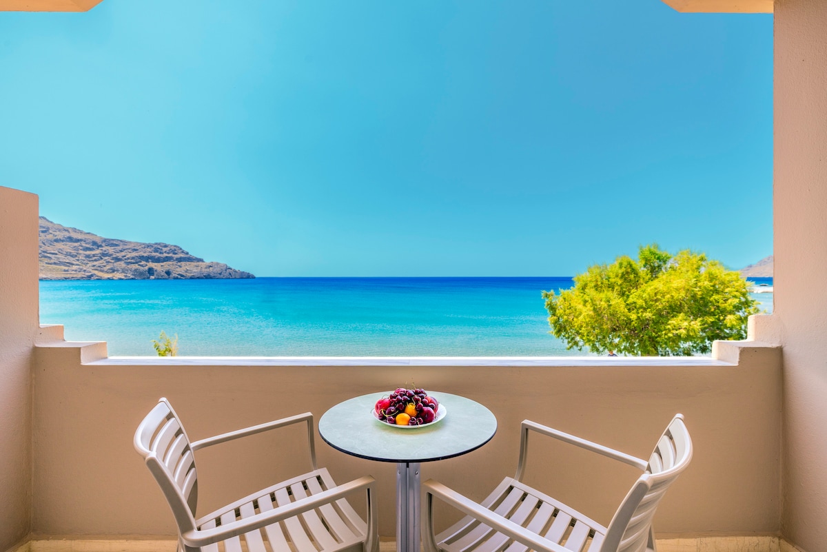 A private balcony offers a table and two chairs, with a bowl of fresh fruits placed centrally. The view overlooks the clear blue waters of the sea, flanked by lush greenery and distant hills under a bright blue sky.