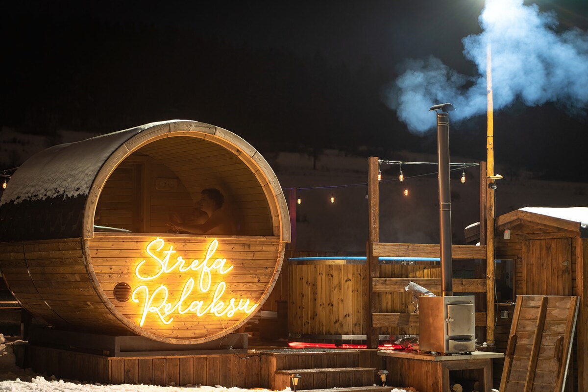 The relaxation area is highlighted at night with a wooden barrel sauna, surrounded by snow. A neon sign reading 'Strefa Relaksu' is visible. A wooden deck with a hot tub and a smokestack emits soft wisps of smoke into the night sky, enhancing the tranquil ambiance.