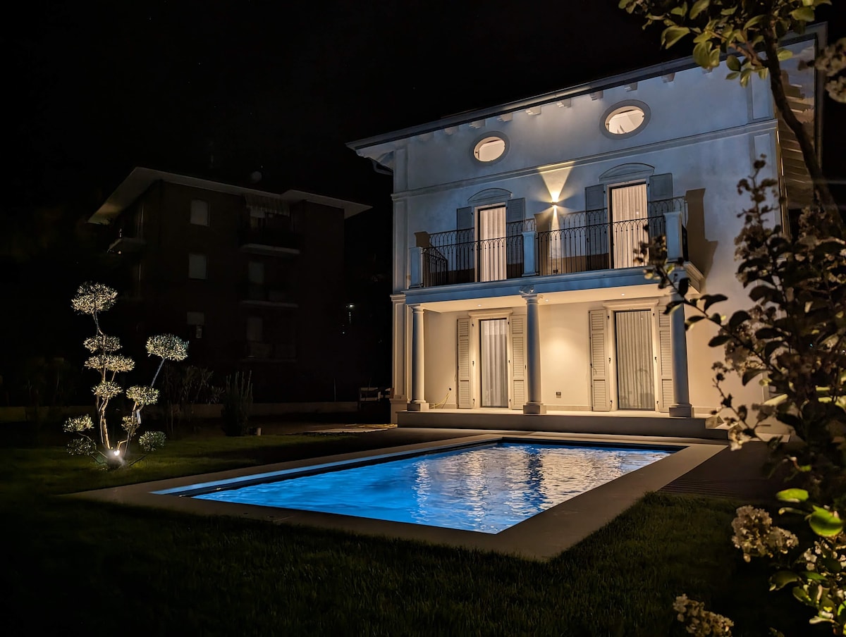 The exterior of a villa is illuminated at night, featuring two balconies with open doors. A well-maintained pool reflects soft blue light, surrounded by neatly trimmed grass. Decorative lighting outlines a plant sculpture, enhancing the yard's visual appeal.