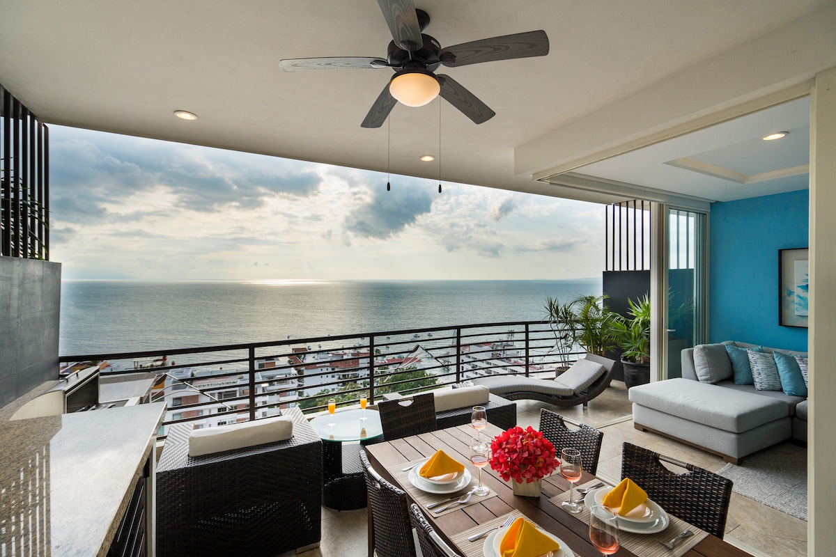 A spacious terrace offers an outdoor dining area with a round table set for a meal, complemented by elegant glassware and vibrant yellow napkins. Comfortable lounge chairs and a modern sectional sofa are arranged, with a panoramic ocean view visible beyond the railing.