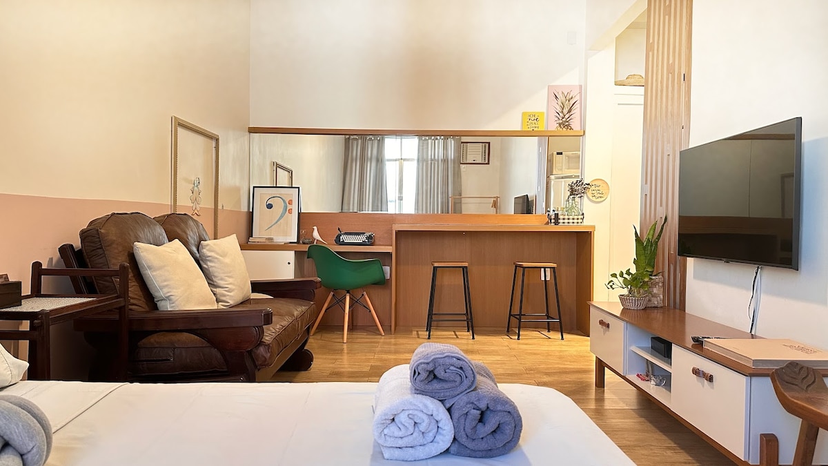A well-furnished studio apartment features a seating area with a brown leather couch, adorned with light-colored cushions. A wooden counter with two stools provides a space for dining or working. A wall-mounted television is positioned near a decorative plant on a low cabinet.