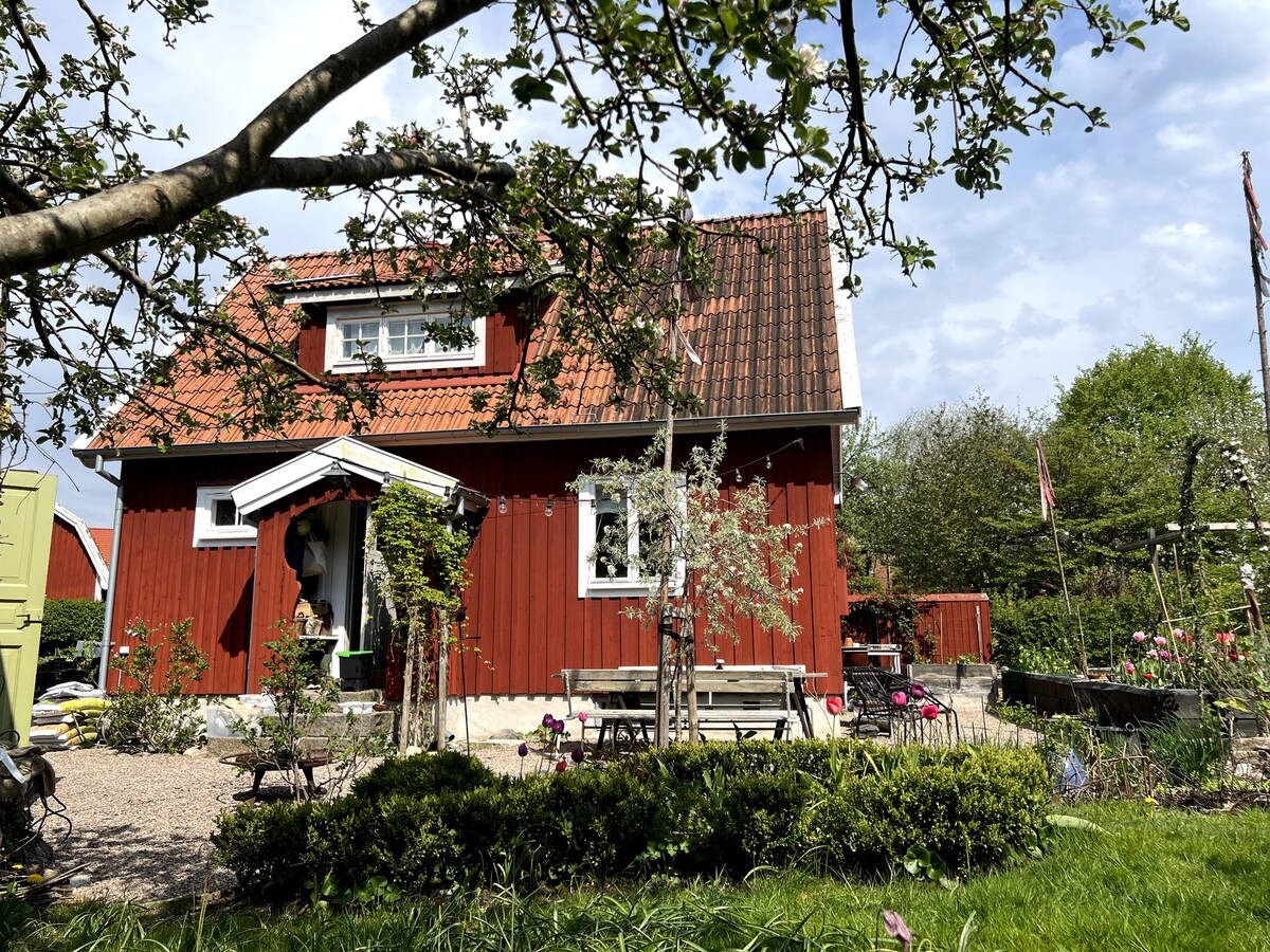 A charming red house is surrounded by lush greenery and colorful flowers, showcasing a welcoming entrance. A bench is positioned in the front garden, and the roof features traditional tiles. The home is set against a partly cloudy sky, enhancing the peaceful ambiance of the outdoor space.