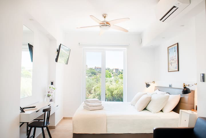 Luminoso segundo dormitorio con una acogedora cama doble, escritorio y silla, ventilador de techo y aire acondicionado, TV de pantalla plana con Netflix y grandes puertas de ventana que se abren a vistas tranquilas de la naturaleza circundante.