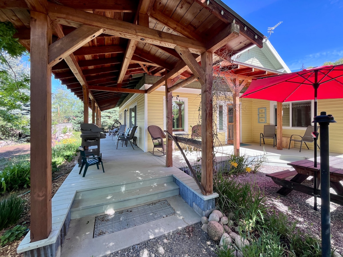 An outdoor patio area features exposed wooden beams and a shaded entrance. Seating options include two chairs and a picnic table, while a grill is positioned nearby. A red umbrella provides additional shade, and colorful flowers surround the space, creating a welcoming environment.