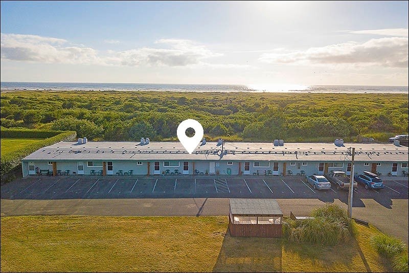 An aerial view of the condo complex is shown, located amidst lush greenery with the ocean visible in the distance. The building features multiple units, each with outdoor seating options. A parking area is situated in front, highlighting accessibility and openness to the natural surroundings.