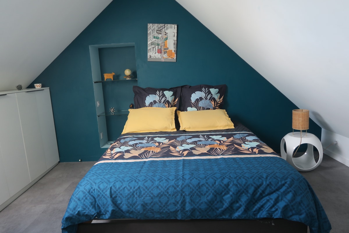 The sleeping area is defined by a spacious bed adorned with patterned bedding in shades of blue and yellow. A decorative shelf with accessories is placed against a teal wall. Natural light enters through a nearby window, while a nightstand and artwork add to the room's character.