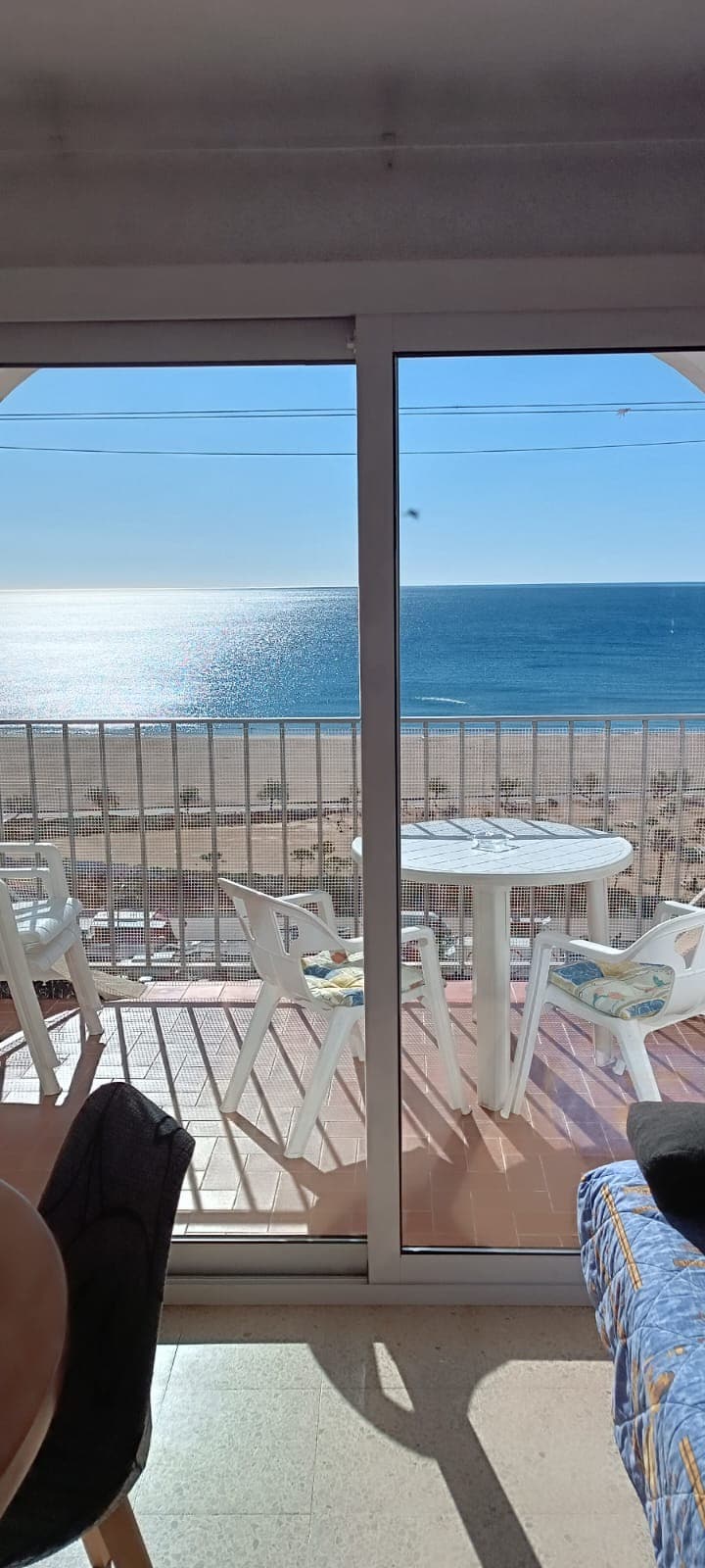 A balcony offers a panoramic view of the beach and the ocean, with sunlight reflecting off the water. A round white table is accompanied by four matching chairs, providing a comfortable space for outdoor relaxation.